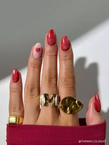 A stylish manicure featuring glittery red nails with a soft pink accent nail adorned with a small red heart.