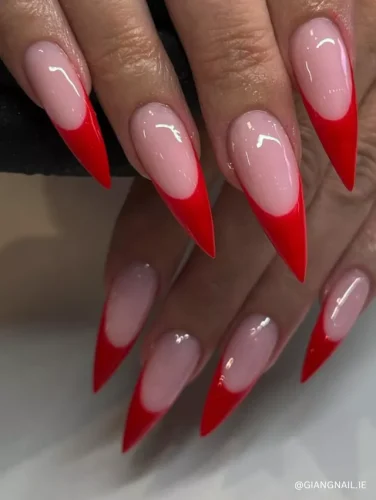 A bold stiletto manicure featuring nude pink nails with dramatic, sharp red French tips.