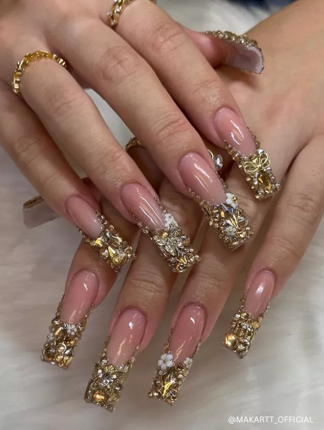 Luxurious nude coffin nails adorned with opulent gold embellishments, rhinestones, and intricate floral details for a regal and glamorous look.