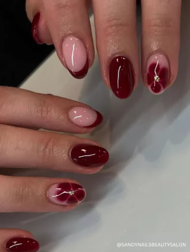 A sophisticated nail design featuring deep red polish, soft red French tips, and delicate floral petal accents with gold embellishments.