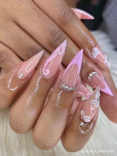 Elegant stiletto nails with soft pink and white swirls, delicate rhinestone accents, and 3D floral details for a chic and feminine look.