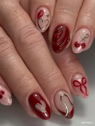 A detailed nail design featuring deep red polish, gold heart outlines, 3D swirls, cherry motifs, and delicate ribbon accents.