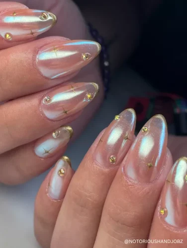 Iridescent almond nails with a pearly chrome finish, delicate gold French tips, star accents, and rhinestone embellishments for a celestial glow.