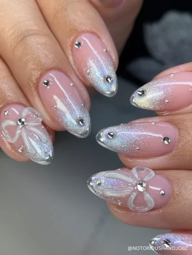 Chic almond nails with a soft pink base, shimmering silver glitter tips, rhinestone accents, and 3D white bow details for a delicate yet glamorous touch.