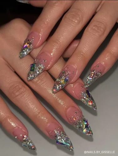 Dazzling stiletto nails with a nude base, covered in silver glitter, iridescent rhinestones, and chunky crystal embellishments for a high-glam look.