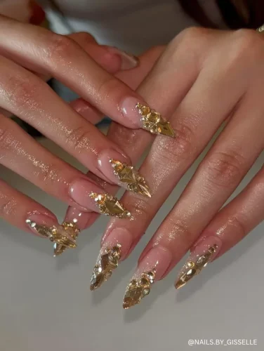 Luxurious stiletto nails with a nude base, adorned with gold rhinestones and oversized crystal embellishments for a regal, high-fashion look.