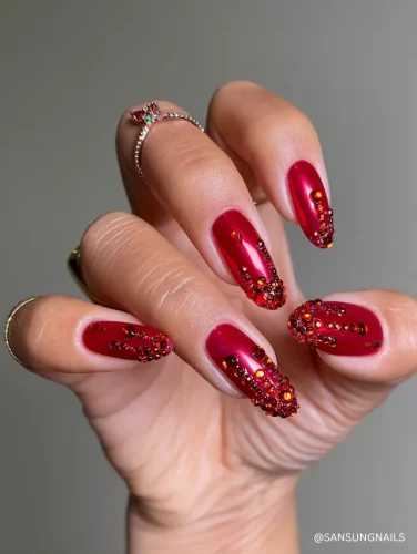 Bold almond nails with a deep ruby red chrome finish, featuring cascading red and black rhinestone embellishments for a fiery, luxurious look.