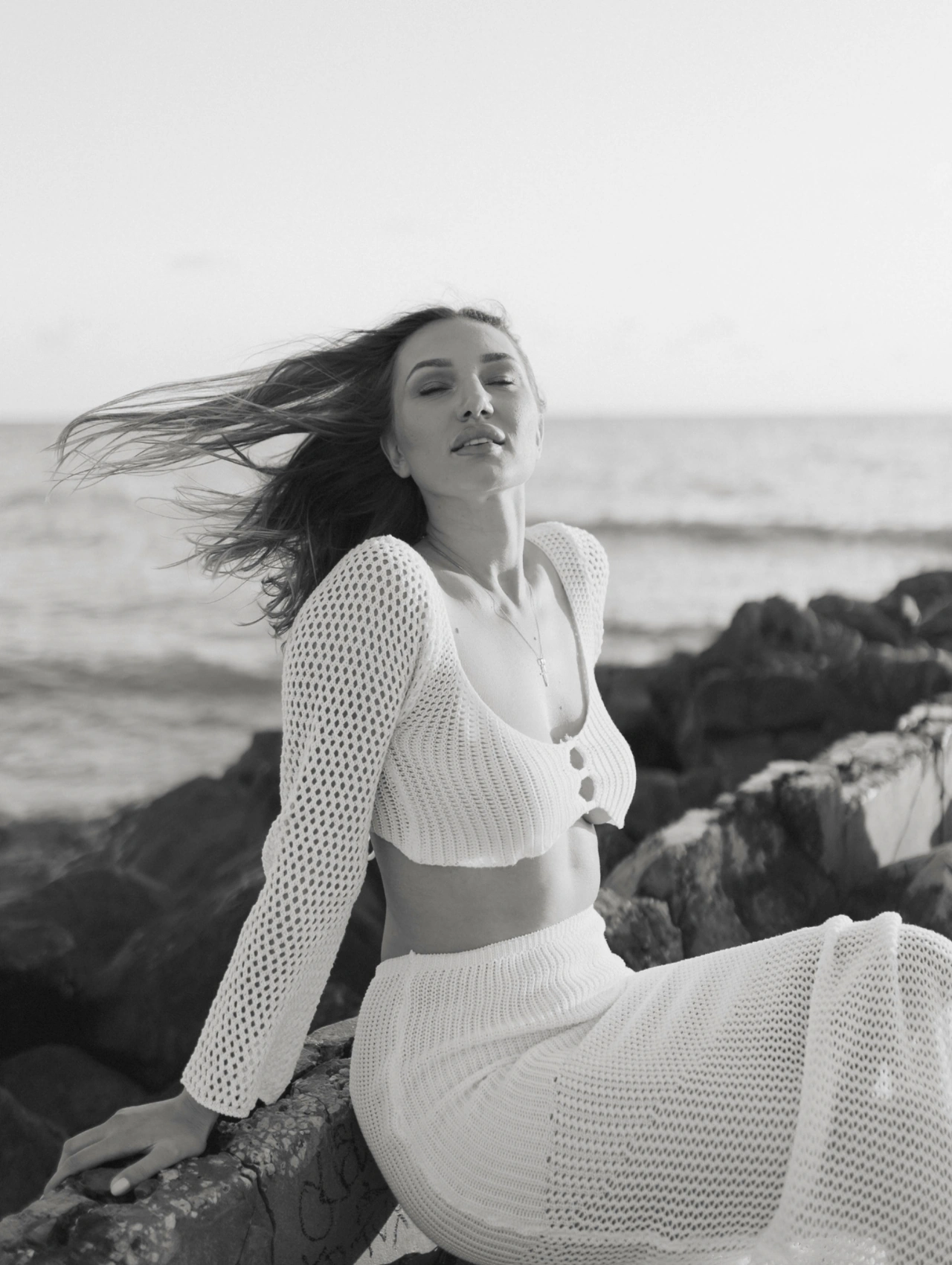 Final Glow-Up_ Own Every Look, Every Moment, black-and-white beach photo of a model sitting on seaside rocks wearing a white crochet set, spring break outfits