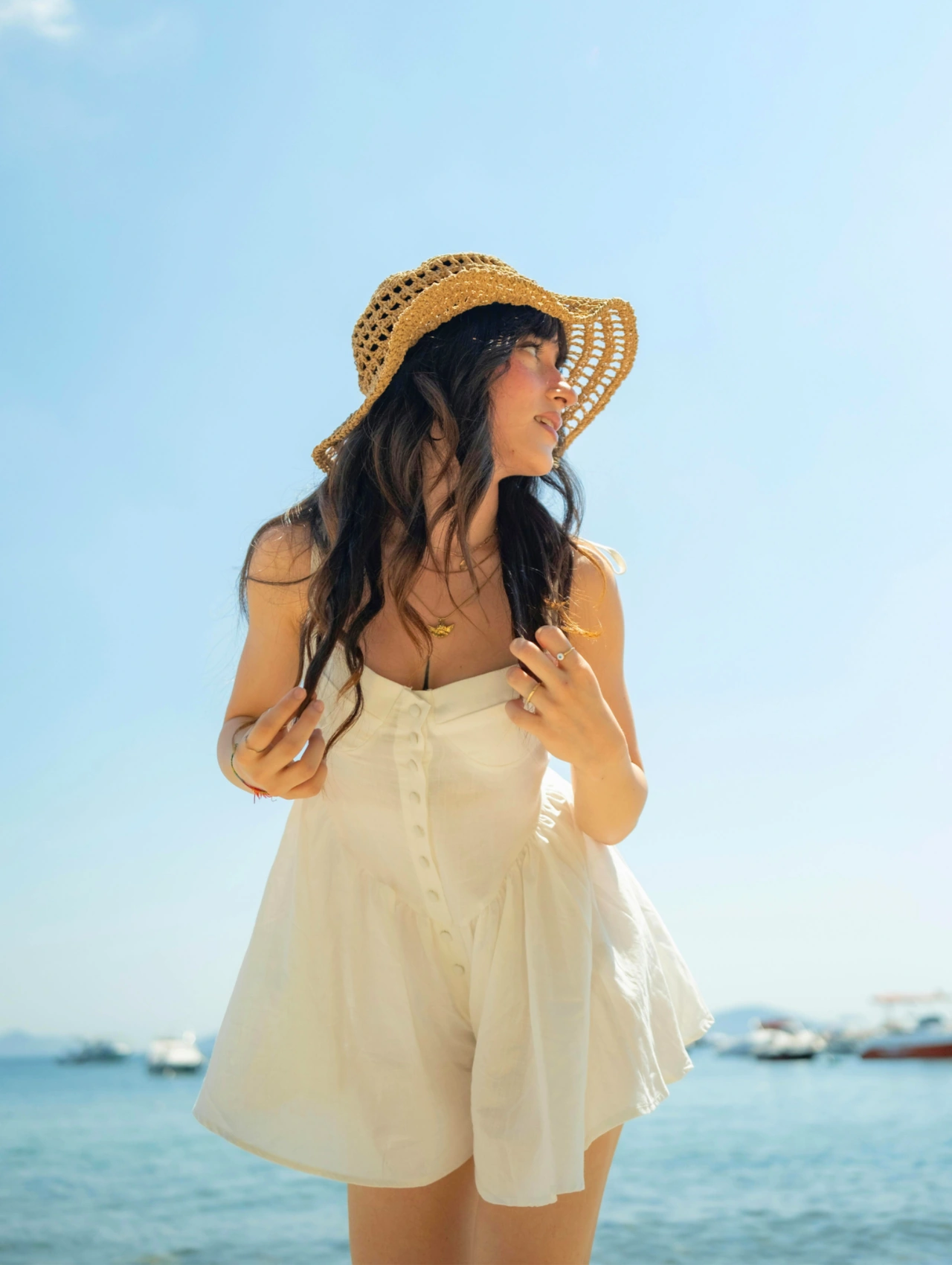 Para finalizar, ready para brillar esta Semana Santa, mujer con vestido blanco ligero y sombrero de paja frente al mar, inspiración de ropa de playa para un look luminoso.