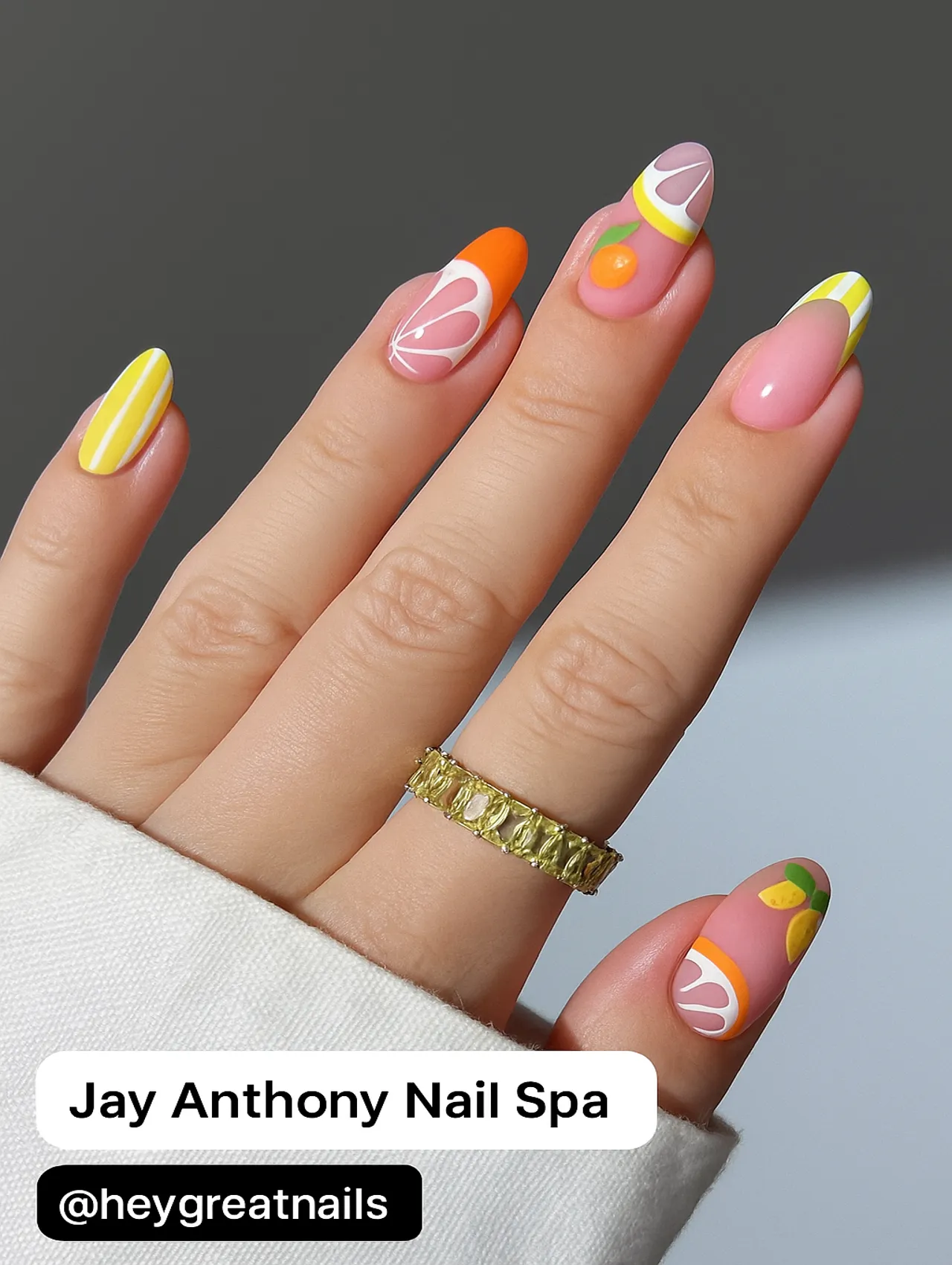 A close-up of a vibrant citrus-themed nail design from Jay Anthony Nail Spa, featuring colorful fruit patterns on almond-shaped nails.