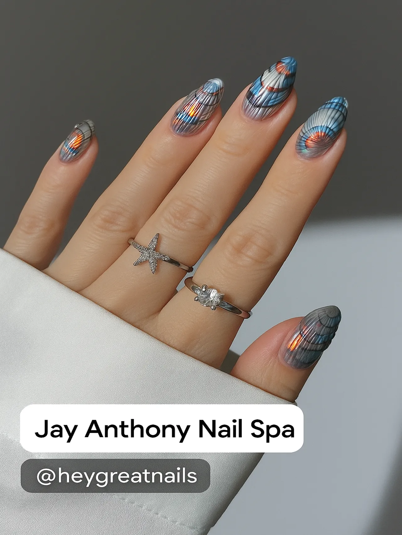 Close-up of a hand with iridescent seashell-patterned nail art, wearing silver rings, with Jay Anthony Nail Spa branding at the bottom.