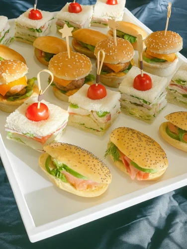 Bandeja blanca con bocas para fiestas que incluye mini sándwiches, sliders con pan de sésamo, y bocadillos con tomate cherry, sobre fondo oscuro.