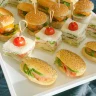 Bandeja blanca con bocas para fiestas que incluye mini sándwiches, sliders con pan de sésamo, y bocadillos con tomate cherry, sobre fondo oscuro.