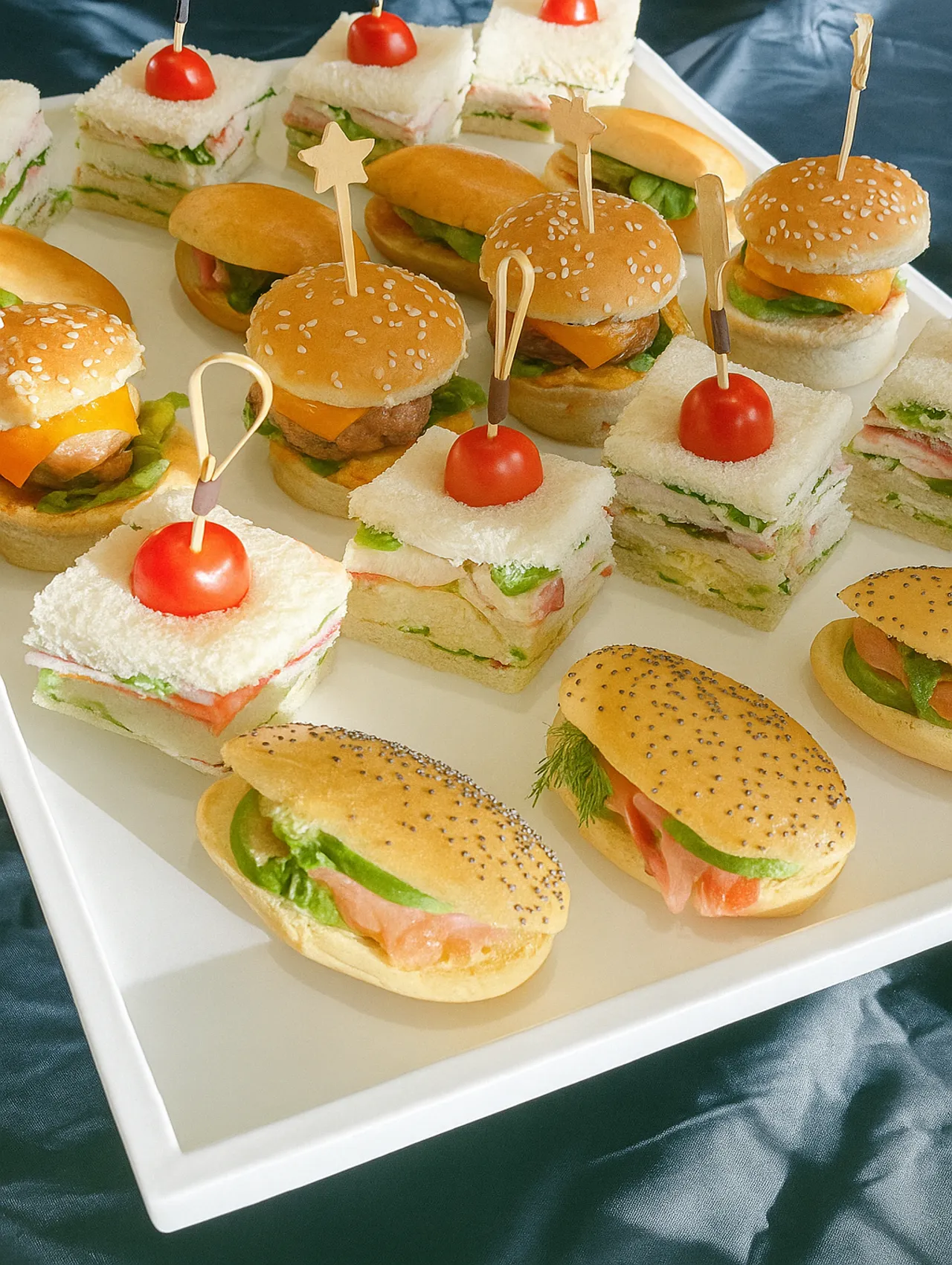 Bandeja blanca con bocas para fiestas que incluye mini sándwiches, sliders con pan de sésamo, y bocadillos con tomate cherry, sobre fondo oscuro.