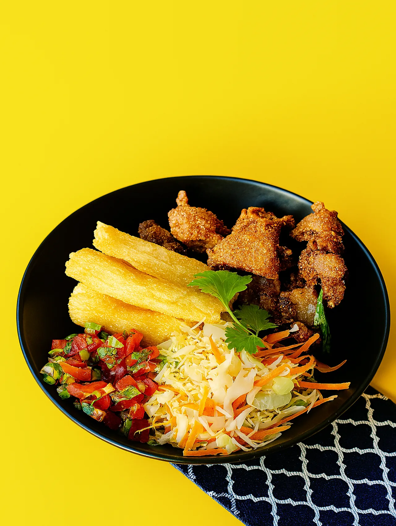Plato con chicharrones crujientes, yuca frita, curtido de repollo y pico de gallo servido en plato negro sobre fondo amarillo.