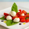 Cucharitas de ensalada caprese con tomate cherry, bolitas de mozzarella y hojas de albahaca sobre plato blanco