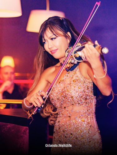 A glamorous female violinist performing live at an upscale Orlando nightlife venue, wearing a sparkling dress under ambient lighting.