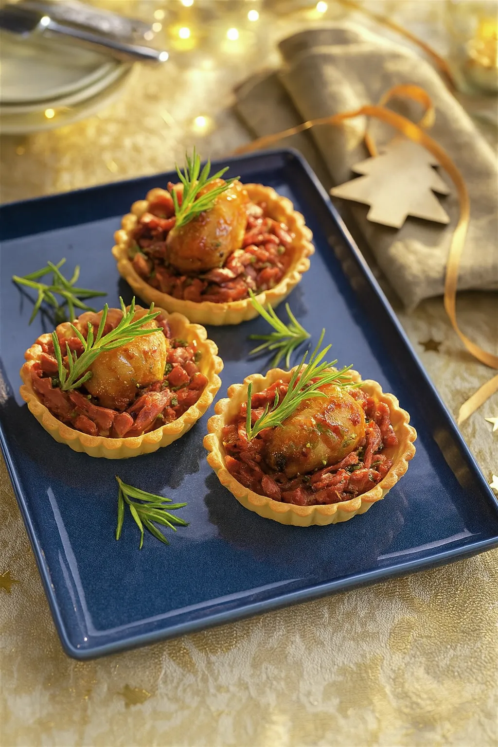 Tartaletas doradas rellenas de atún con mayonesa y verduras, decoradas con papas caramelizadas y romero fresco sobre un plato azul.
