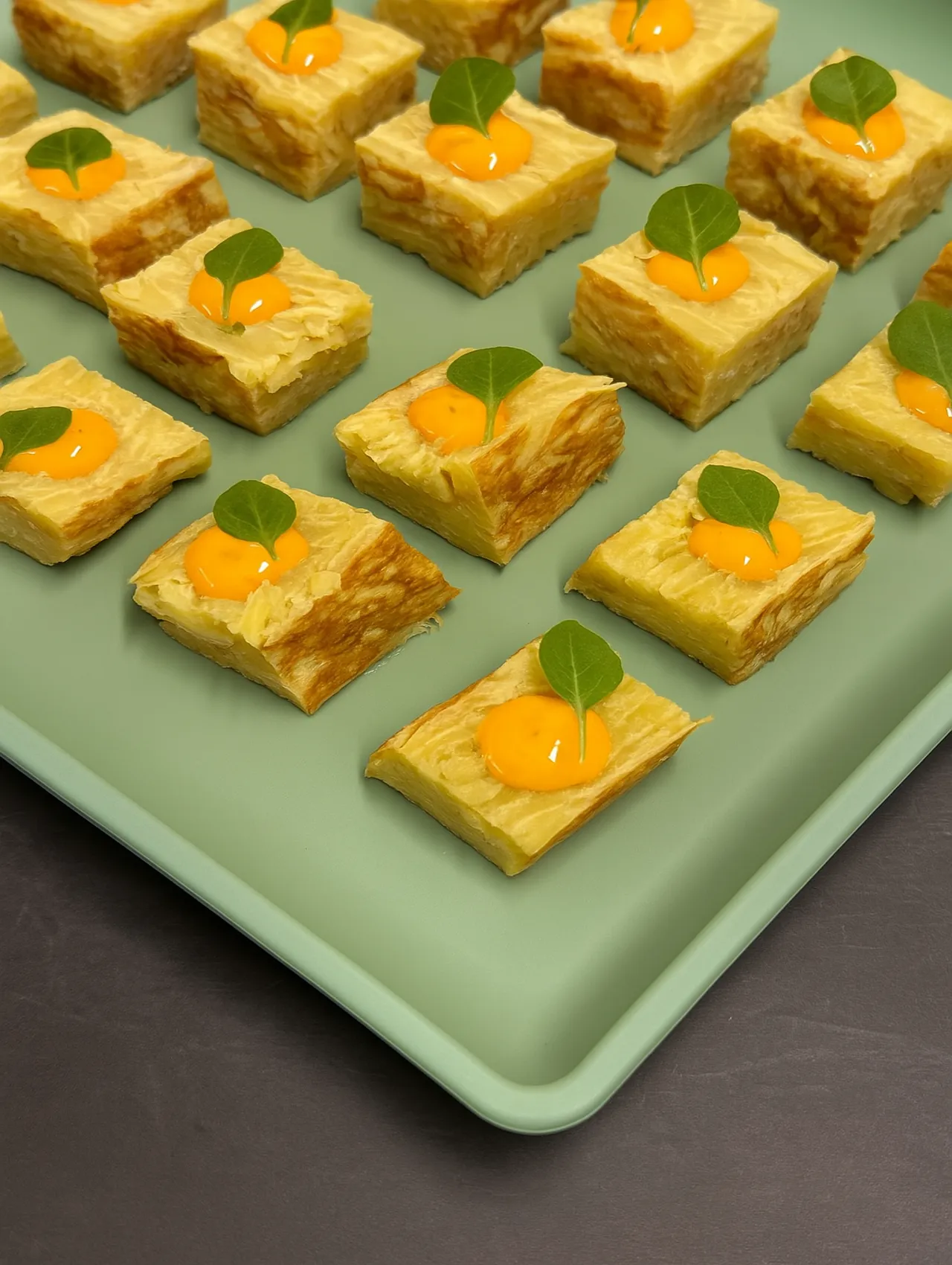 Cubitos de tortilla española con un toque de alioli y hoja verde sobre bandeja verde.