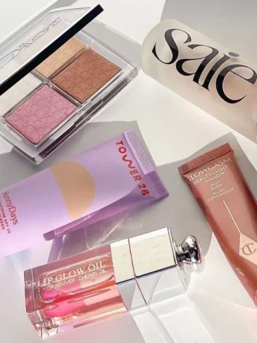 Flat lay of luxury and trending beauty products including Dior Lip Glow Oil, Charlotte Tilbury Glowgasm, Tower 28 blush, Saie foundation, and an eyeshadow palette in neutral and pink shades.