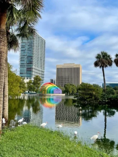Scenic view of Lake Eola in downtown Orlando, Florida, featuring the iconic rainbow bandshell, modern high-rise buildings, palm trees, and white ibis birds along the water's edge.