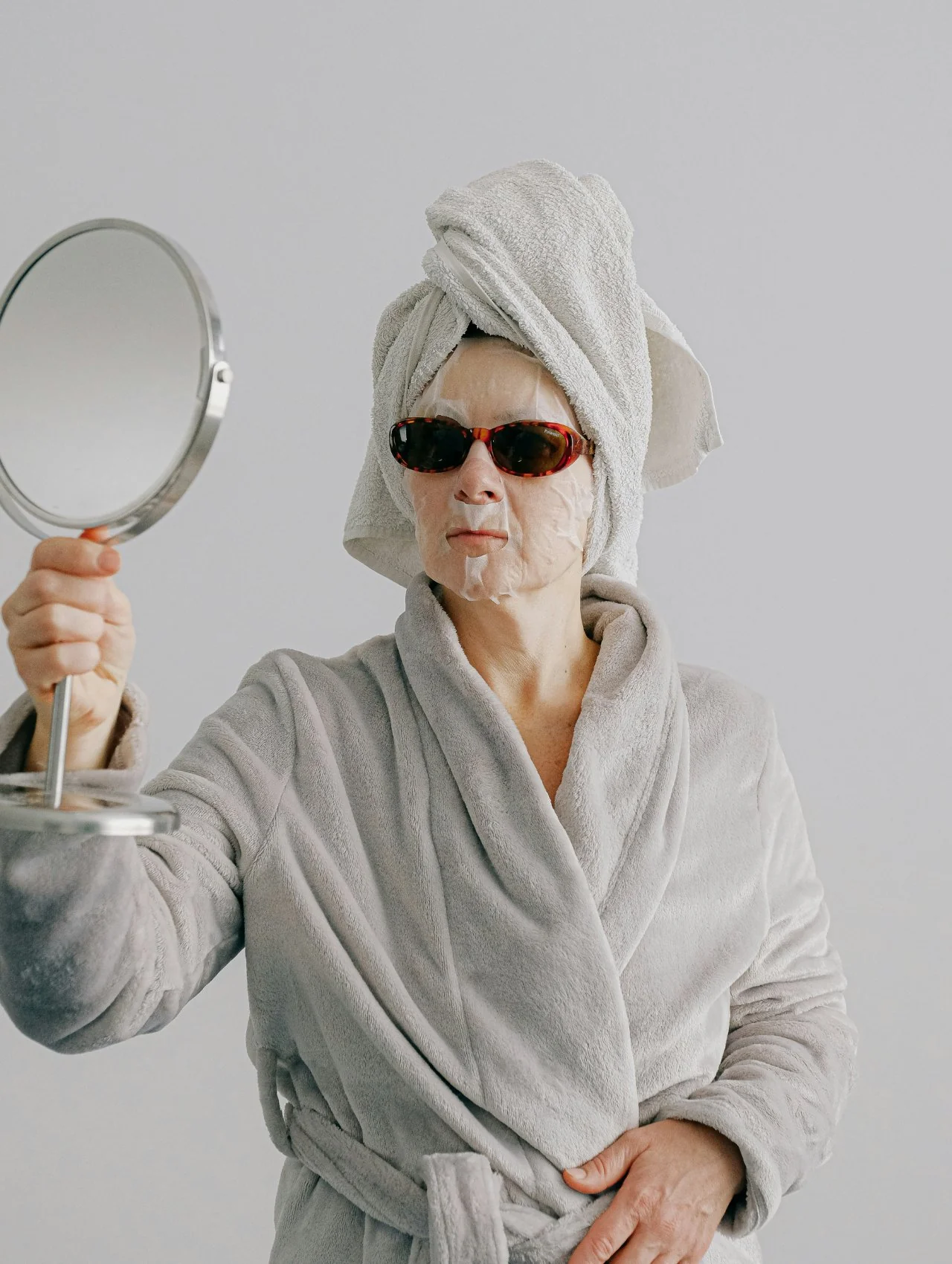Woman in robe and sheet mask checking a handheld mirror, illustrating common triggers to avoid in the best skincare for sensitive skin