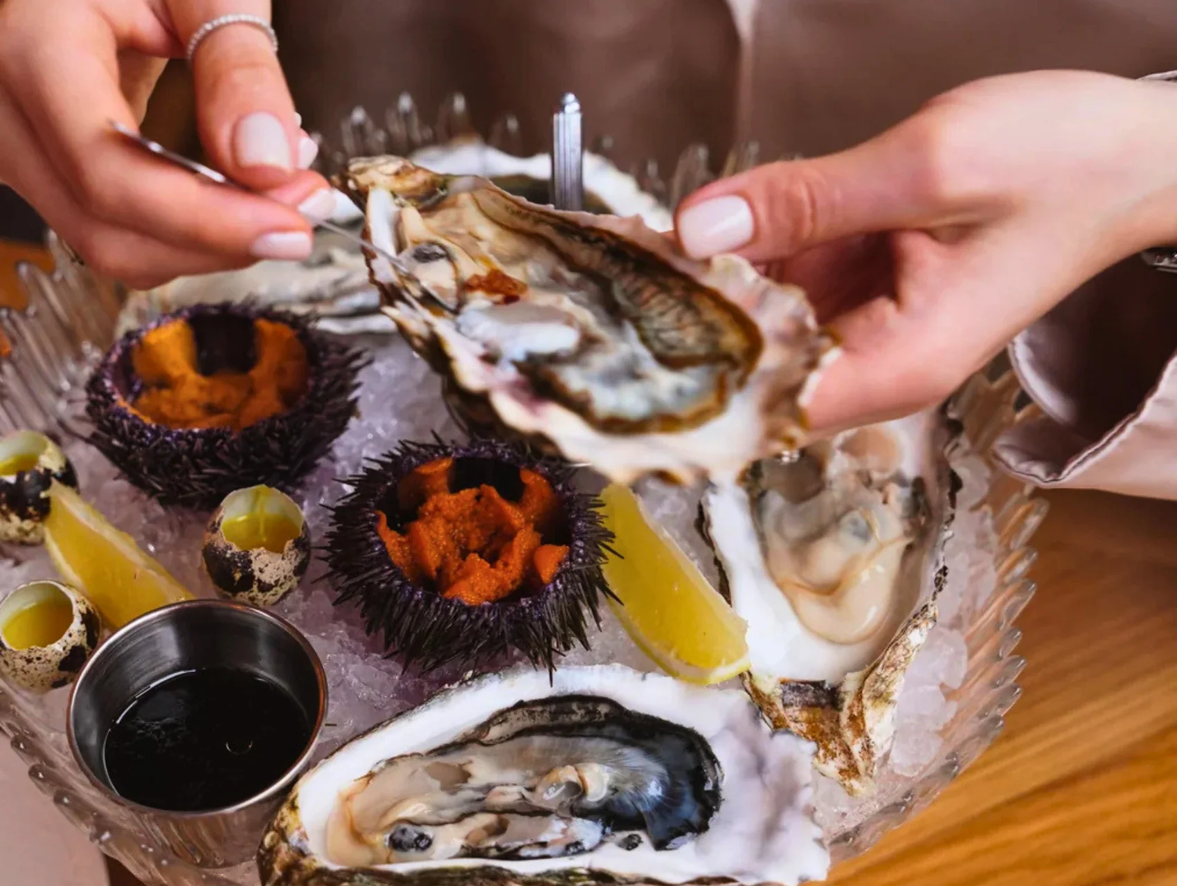 Hands lifting an oyster from an iced platter with sea urchin and lemon, opening a delicious gourmet meal for two